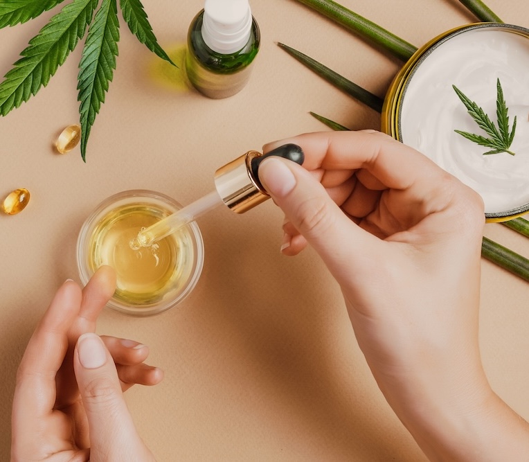 Pipette with CBD cosmetic oil in female hands on a table background with cosmetics, cream with cannabis and hemp leaves.